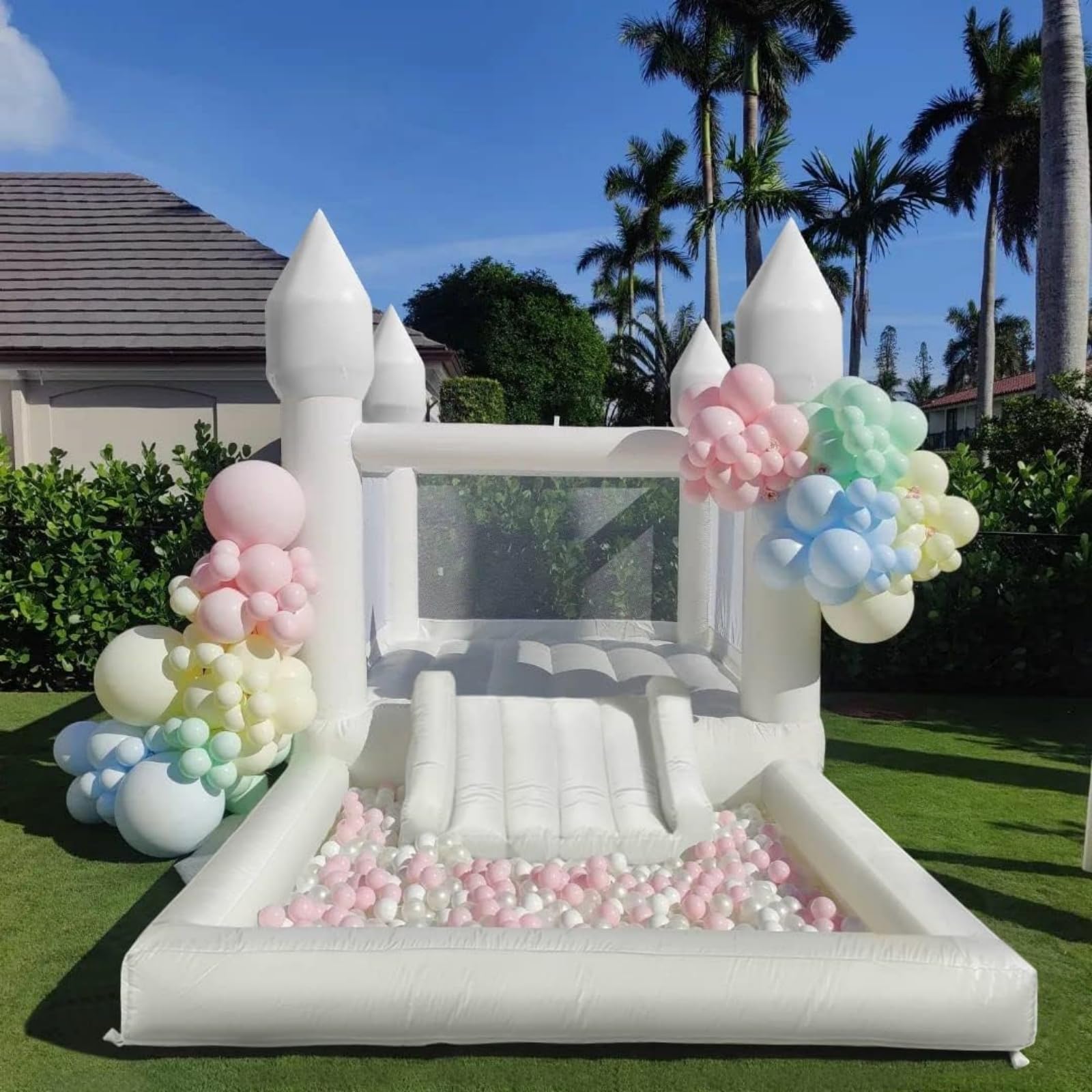 White inflatable bounce house with a slide and pastel balloon decorations, set on green grass under a blue sky.