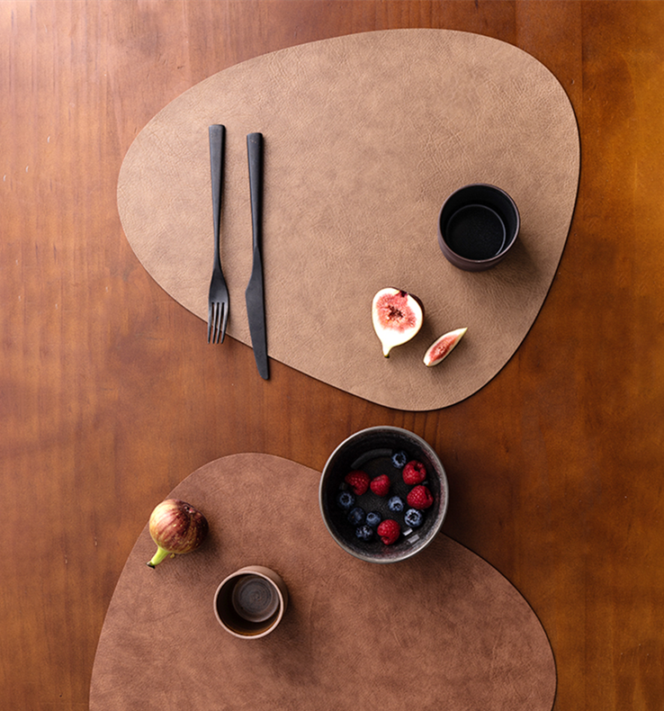 Two waterproof leather placemats in light brown and dark brown are set on a wooden table, accompanied by black cutlery and bowls of fruit.