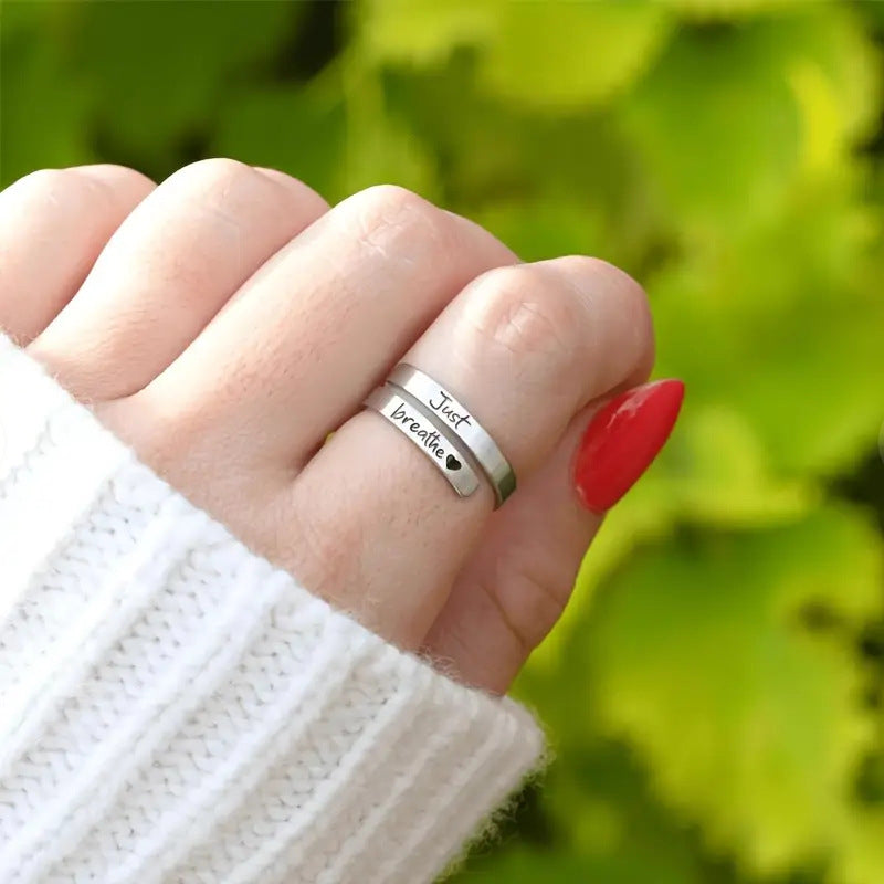 Silver heart-shaped spiral ring sits on a hand with red nails, featuring "Just Breathe" engraving. Worn with a cozy white sweater.